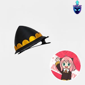 May include: Black hair clip with a gold scalloped trim, resembling a hat. The clip is positioned on a white background, with a cartoon image of a character with pink hair and the word "ANYA" visible.