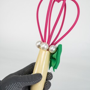 K&ouml;nnte beinhalten: Ein dekorativer Schneebesen mit einem hellgelben Griff, verziert mit drei Kunstperlen und einer gr&uuml;nen Schleife. Der Schneebesen hat leuchtend rosa Schlaufen, die ein skurriles Design ergeben. Das Objekt wird von einer behandschuhten Hand gehalten.