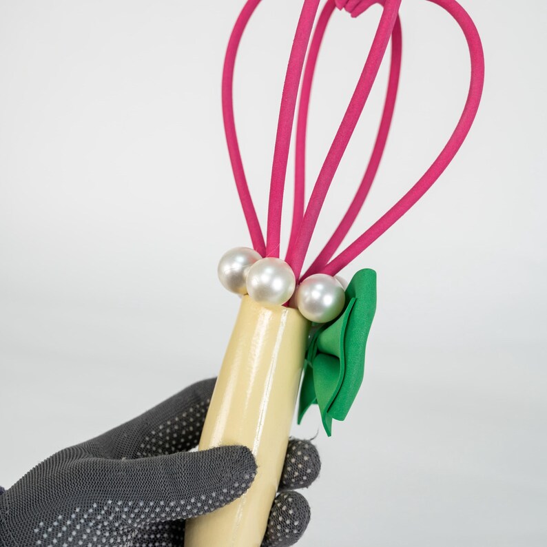 K&ouml;nnte beinhalten: Ein dekorativer Schneebesen mit einem hellgelben Griff, verziert mit drei Kunstperlen und einer gr&uuml;nen Schleife. Der Schneebesen hat leuchtend rosa Schlaufen, die ein skurriles Design ergeben. Das Objekt wird von einer behandschuhten Hand gehalten.