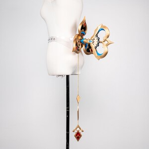 May include: An elaborate gold-coloured accessory with red and blue details, attached to a clear belt. The design features a stylized butterfly shape with a dangling chain ending in a red jewel. The accessory is displayed on a white mannequin.