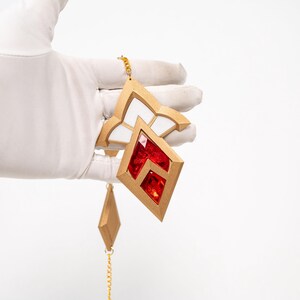 May include: Geometric earrings in gold with red and white details. The earrings feature a diamond shape with a red centre and a gold frame. A smaller diamond hangs below on a gold chain. The earrings are held by a gloved hand.