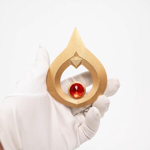 May include: A gold-coloured pendant in a teardrop shape, with a red gemstone at its centre. The pendant has a pointed top and a star-shaped design element. The image is set against a white background.