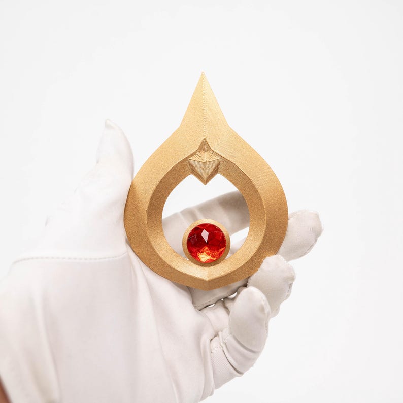 May include: A gold-coloured pendant in a teardrop shape, with a red gemstone at its centre. The pendant has a pointed top and a star-shaped design element. The image is set against a white background.