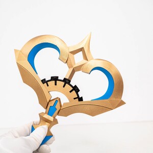 May include: A gold-coloured, stylised key with blue and black accents. The key features a gear-like design in the centre and a heart-shaped top. The handle is held by a gloved hand.