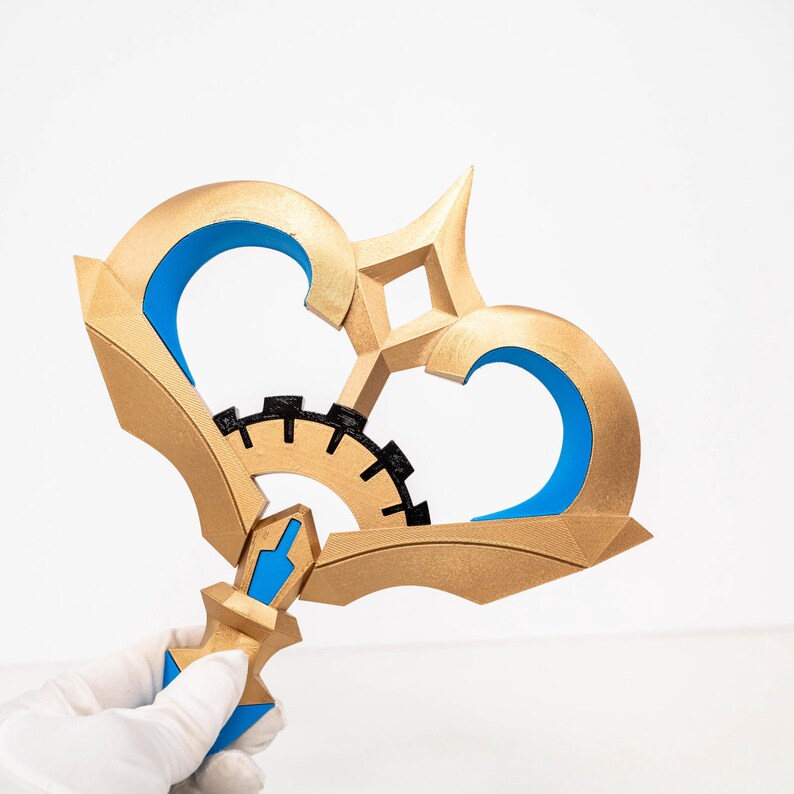 May include: A gold-coloured, stylised key with blue and black accents. The key features a gear-like design in the centre and a heart-shaped top. The handle is held by a gloved hand.