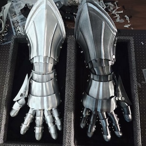 May include: Two metal gauntlets, one silver and one dark gray, are displayed side-by-side. The gauntlets are articulated, with individual finger segments and protective plates. The design suggests medieval armor, with a focus on protection and a detailed, layered construction.