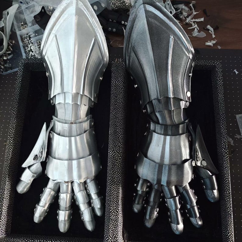 May include: Two metal gauntlets, one silver and one dark gray, are displayed side-by-side. The gauntlets are articulated, with individual finger segments and protective plates. The design suggests medieval armor, with a focus on protection and a detailed, layered construction.