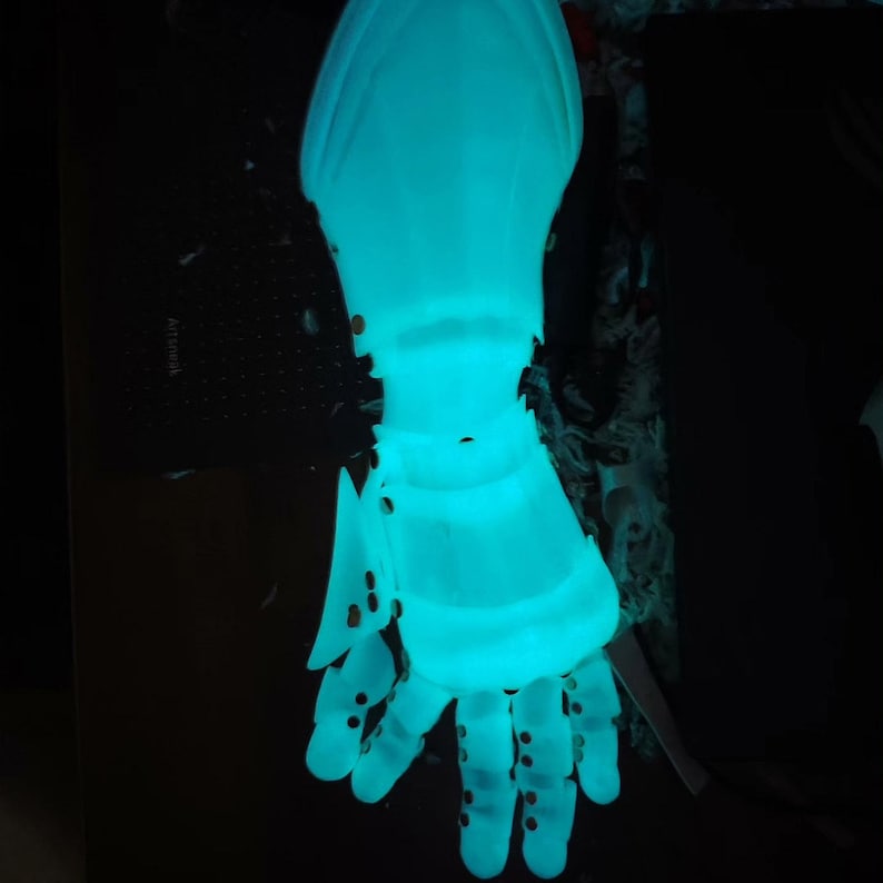 May include: A glowing, light blue articulated hand, possibly a prop or sculpture. The hand is detailed with visible joints and small circular perforations. The overall effect is futuristic and ethereal, with a strong visual impact against the dark background.