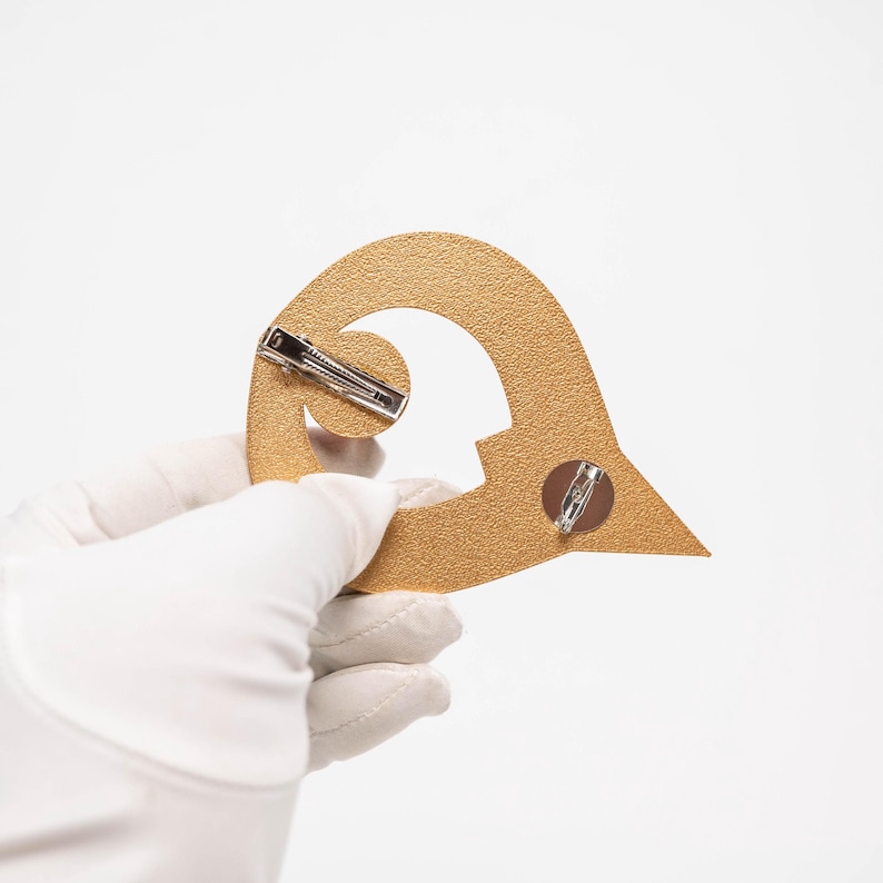 May include: Gold-coloured, textured metal pin in a stylised shape, possibly a logo or emblem. The pin has two silver-coloured clasps on the back for attachment. The pin is held by a gloved hand against a white background.