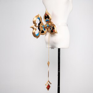 May include: An elaborate gold-coloured accessory with blue and black detailing, featuring a central design and a dangling chain with red and gold geometric shapes. The accessory is displayed on a white mannequin.