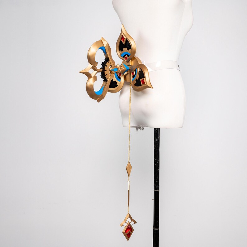 May include: An elaborate gold-coloured accessory with blue and black detailing, featuring a central design and a dangling chain with red and gold geometric shapes. The accessory is displayed on a white mannequin.