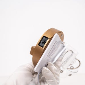 May include: A gold-coloured, circular object with a black button and a white rectangular plate attached. The plate holds a clear, curved plastic piece with metal accents. The object is held by a gloved hand.