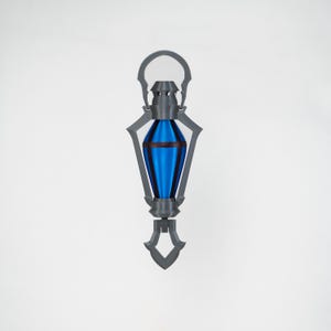 May include: A grey and blue decorative lantern with a handle at the top and a decorative base. The lantern has a blue, teardrop-shaped centre with a dark red band. The lantern is designed with a geometric, art deco style.