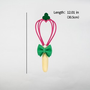 K&ouml;nnte beinhalten: Ein skurriler Zauberstab mit einem herzf&ouml;rmigen rosa Oberteil, einer gr&uuml;nen Schleife und einem Perlenakzent. Der Zauberstab ist 30,5 cm lang. Der Griff ist hellgelb.
