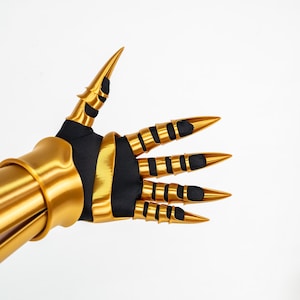 Puede incluir: Un guantelete articulado de color dorado con una base de tela negra, placas met&aacute;licas doradas para los dedos y puntas en forma de garra. El dise&ntilde;o incluye un brazalete dorado s&oacute;lido y piezas de armadura segmentadas que cubren cada dedo, creando una apariencia afilada y con garras con un acabado suave y reflectante en los componentes dorados.