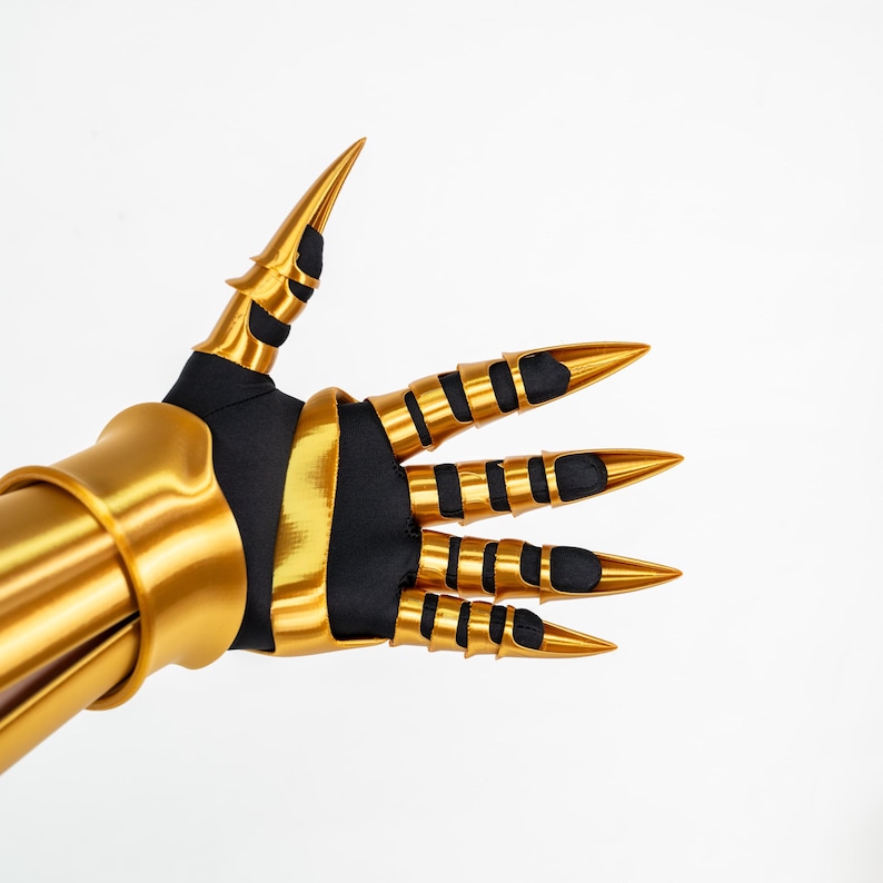 Puede incluir: Un guantelete articulado de color dorado con una base de tela negra, placas met&aacute;licas doradas para los dedos y puntas en forma de garra. El dise&ntilde;o incluye un brazalete dorado s&oacute;lido y piezas de armadura segmentadas que cubren cada dedo, creando una apariencia afilada y con garras con un acabado suave y reflectante en los componentes dorados.