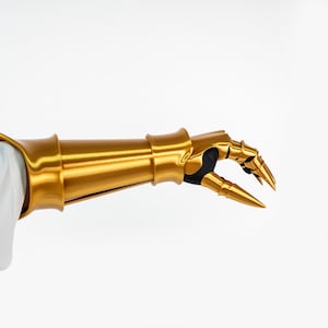 Puede incluir: Un guantelete de metal dorado con una secci&oacute;n de antebrazo cil&iacute;ndrica y lisa, y placas articuladas en los dedos que terminan en puntas afiladas. El interior de la mano y las articulaciones de los dedos est&aacute;n forrados con material negro.