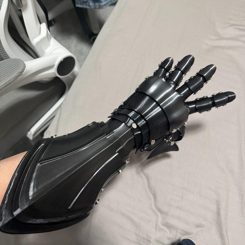 May include: Black articulated gauntlet with a segmented design. The gauntlet features multiple layers of armor plating, riveted together. The fingers are also articulated, allowing for movement. A fantasy-themed accessory.