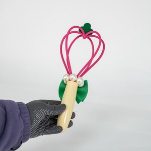 K&ouml;nnte beinhalten: Ein rosa herzf&ouml;rmiger Zauberstab mit einer gr&uuml;nen Schleife und drei wei&szlig;en Perlen. Der Zauberstab hat einen cremefarbenen Griff und einen gr&uuml;nen Akzent oben. Der Zauberstab wird von einer Person mit einem grauen Handschuh gehalten.