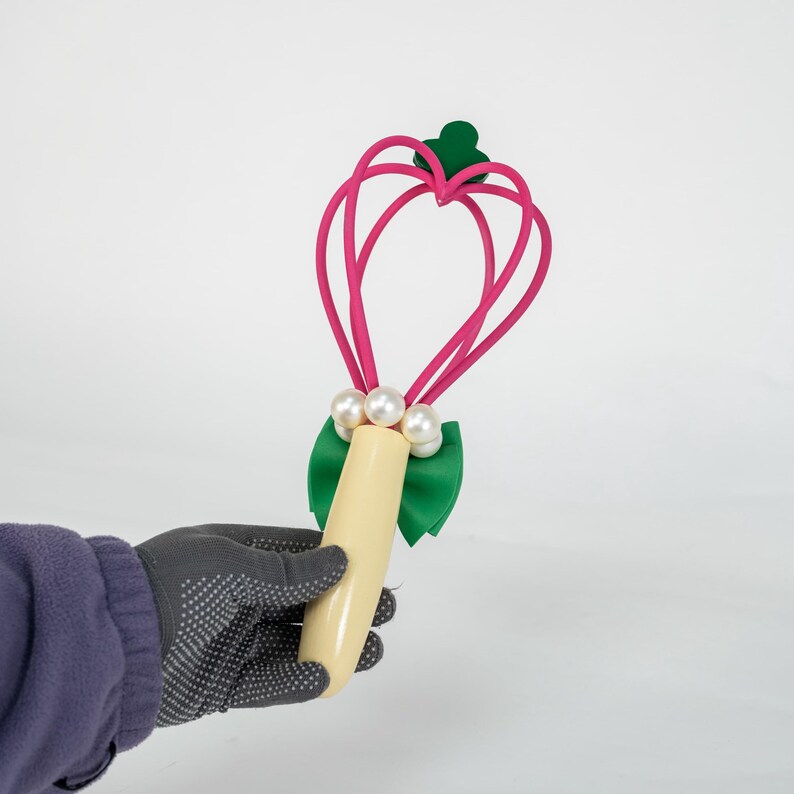 K&ouml;nnte beinhalten: Ein rosa herzf&ouml;rmiger Zauberstab mit einer gr&uuml;nen Schleife und drei wei&szlig;en Perlen. Der Zauberstab hat einen cremefarbenen Griff und einen gr&uuml;nen Akzent oben. Der Zauberstab wird von einer Person mit einem grauen Handschuh gehalten.