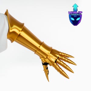 May include: A metallic gold gauntlet featuring a flared forearm guard and articulated finger plates ending in sharp, pointed tips. The piece includes a black fabric base and displays the logo Level Up Persona with an upward arrow icon.