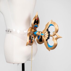 May include: An elaborate gold-coloured accessory with blue and black detailing, attached to a clear belt. The design incorporates geometric shapes and red jewel-like elements. A gold chain hangs from the accessory. The item is displayed on a mannequin.