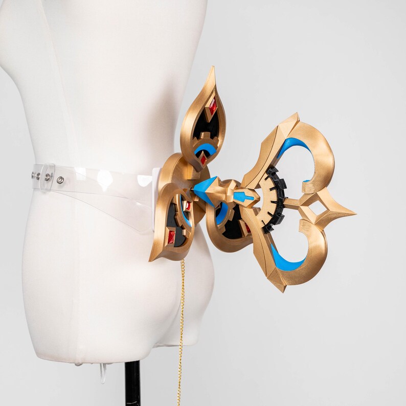May include: An elaborate gold-coloured accessory with blue and black detailing, attached to a clear belt. The design incorporates geometric shapes and red jewel-like elements. A gold chain hangs from the accessory. The item is displayed on a mannequin.