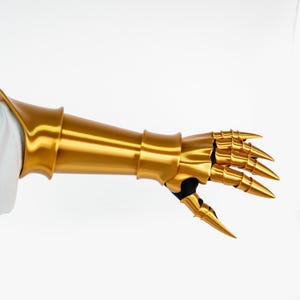 Puede incluir: Un guantelete de metal dorado con una secci&oacute;n de antebrazo lisa y placas de dedos articuladas que se extienden en puntas afiladas. La pieza de armadura est&aacute; construida con placas segmentadas sobre una base negra, cubriendo la mano y el antebrazo.