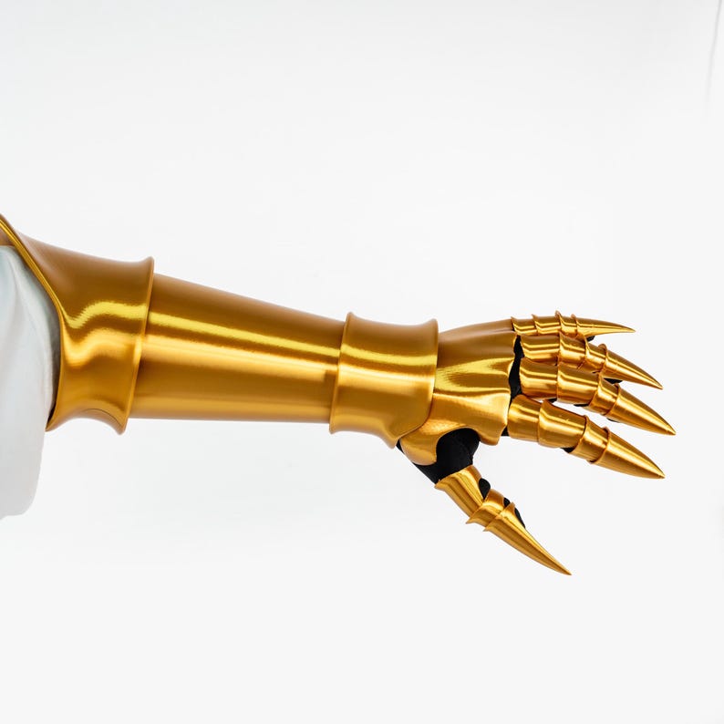 Puede incluir: Un guantelete de metal dorado con una secci&oacute;n de antebrazo lisa y placas de dedos articuladas que se extienden en puntas afiladas. La pieza de armadura est&aacute; construida con placas segmentadas sobre una base negra, cubriendo la mano y el antebrazo.