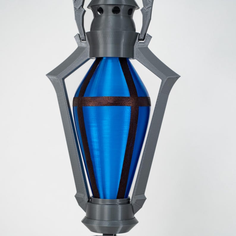 May include: A 3D-printed object with a blue, teardrop-shaped core. The core is accented with dark brown bands and encased in a grey, geometric frame. The top features small holes, and the base is a circular shape. The object has a futuristic design.