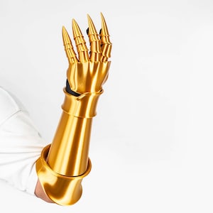 Puede incluir: Un guantelete articulado de color oro met&aacute;lico con una secci&oacute;n de antebrazo lisa y piezas segmentadas para los dedos que terminan en puntas afiladas. Esta pieza de armadura cubre la mano y el antebrazo con un acabado pulido.