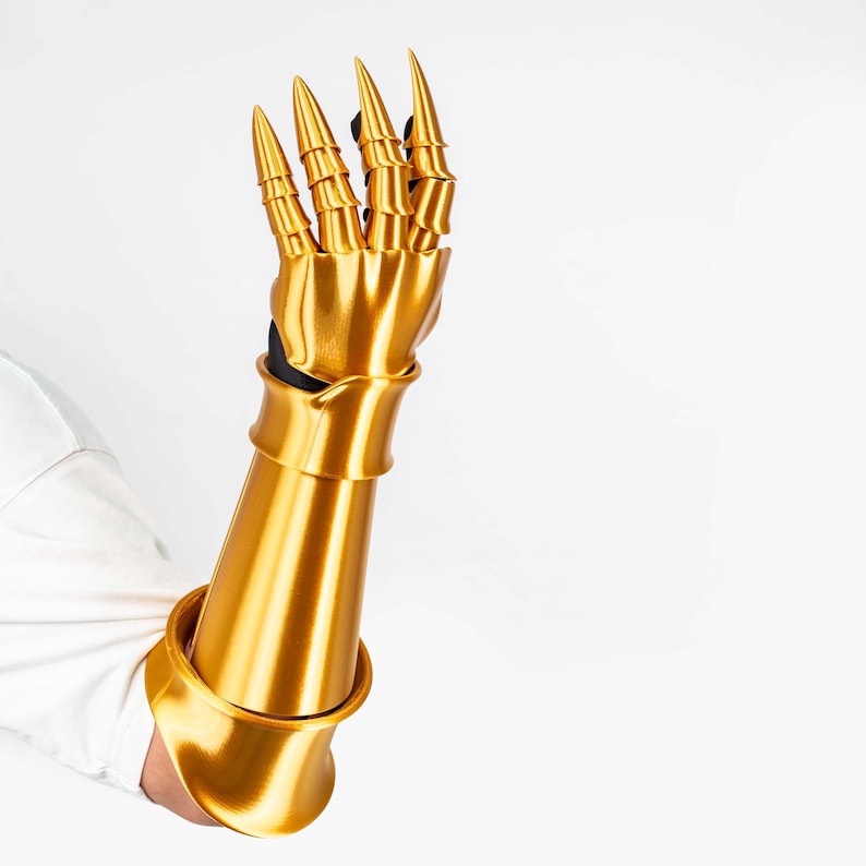 Puede incluir: Un guantelete articulado de color oro met&aacute;lico con una secci&oacute;n de antebrazo lisa y piezas segmentadas para los dedos que terminan en puntas afiladas. Esta pieza de armadura cubre la mano y el antebrazo con un acabado pulido.