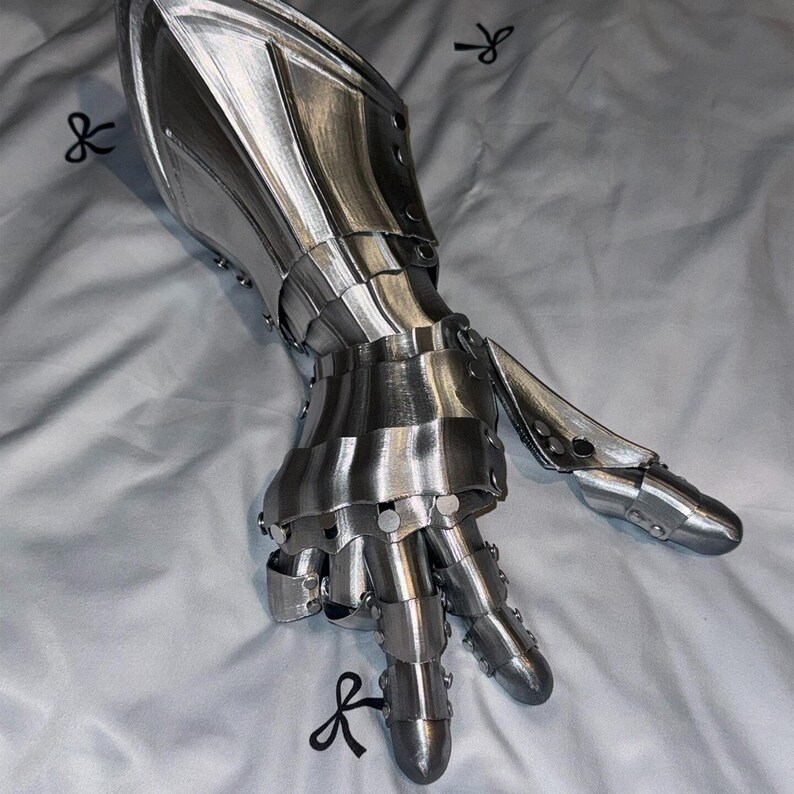 May include: A silver-colored, articulated gauntlet, likely for cosplay or display. The metal hand armor features individual finger segments and a protective forearm piece, all connected with rivets. The surface has a reflective sheen.