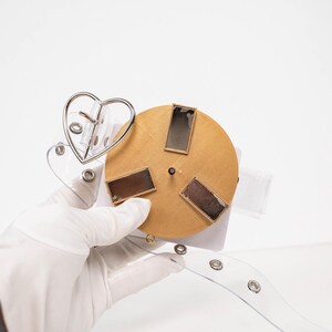 May include: A clear belt with a silver heart-shaped buckle and a gold circular design with three rectangular accents. The belt is held by a gloved hand.
