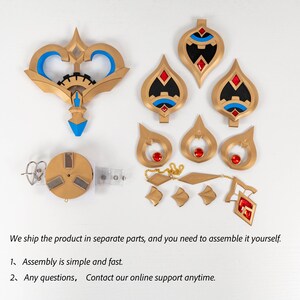 May include: A set of gold-coloured costume pieces, including a large key-shaped item with blue accents, several teardrop-shaped pendants with red gems, and a circular component. The pieces are arranged on a white surface.