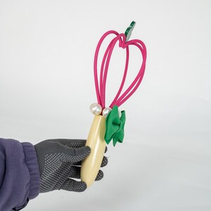 K&ouml;nnte beinhalten: Ein dekoratives Haaraccessoire mit hellgelbem Holzsockel, verziert mit einer gr&uuml;nen Schleife und zwei Kunstperlen. Die Oberseite hat ein rosa herzf&ouml;rmiges Design mit einem gr&uuml;nen Bl&auml;tterakzent.