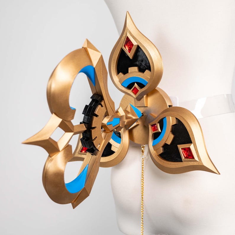 May include: An elaborate gold-coloured accessory with blue and black detailing, and red jewel accents. The design includes a central circular element and a decorative chain. The accessory is attached to a clear strap.
