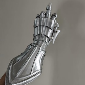 May include: A silver-colored articulated gauntlet, designed to cover the hand and forearm. The gauntlet is made of overlapping metal plates, with rivets and a pointed extension at the wrist. The fingers are also articulated.