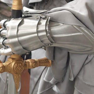 May include: Close-up of a silver metal gauntlet and a gold-colored sword hilt. The gauntlet is articulated with overlapping plates, and the sword hilt features intricate detailing. The background is a soft, gray fabric, suggesting a medieval or fantasy theme.