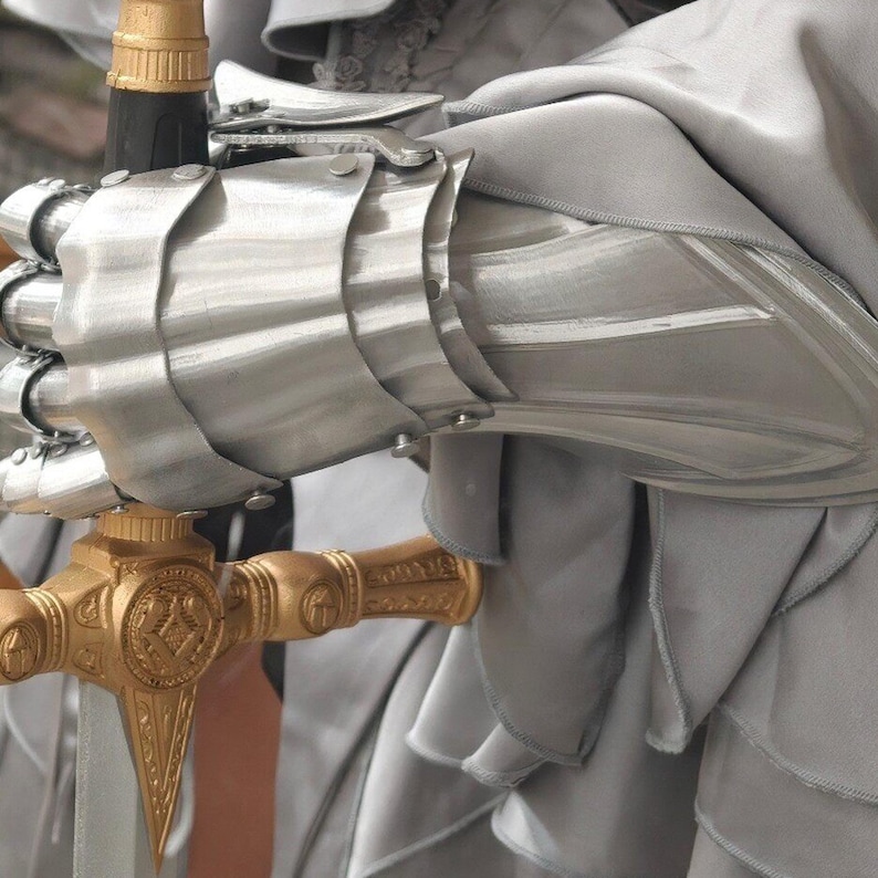 May include: Close-up of a silver metal gauntlet and a gold-colored sword hilt. The gauntlet is articulated with overlapping plates, and the sword hilt features intricate detailing. The background is a soft, gray fabric, suggesting a medieval or fantasy theme.