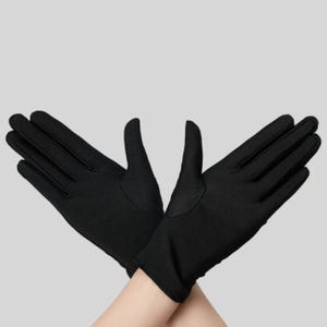 Puede incluir: Un par de guantes negros lisos confeccionados en un tejido opaco y suave. Los guantes est&aacute;n colocados en una posici&oacute;n cruzada, mostrando la longitud total de los dedos y la zona de la mu&ntilde;eca con costuras limpias y sencillas en los bordes.