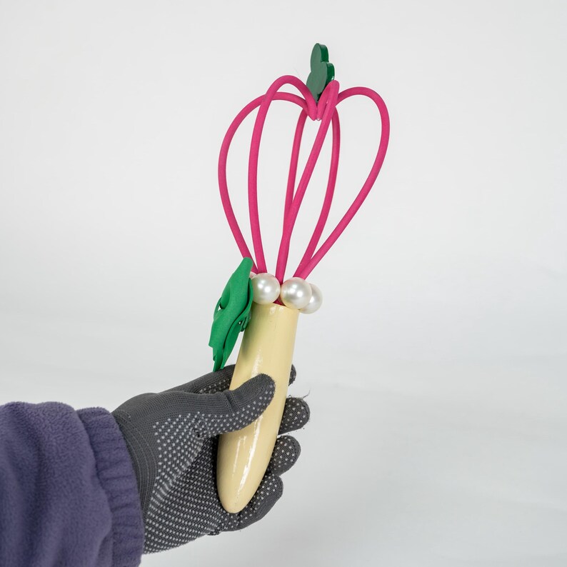 K&ouml;nnte beinhalten: Ein dekorativer K&uuml;chenquirl mit hellgelbem Griff, rosa Drahtschleifen, gr&uuml;nen Blattakzenten und Kunstperlen. Der Schneebesen wird von einer Person mit einem grauen Handschuh gehalten.