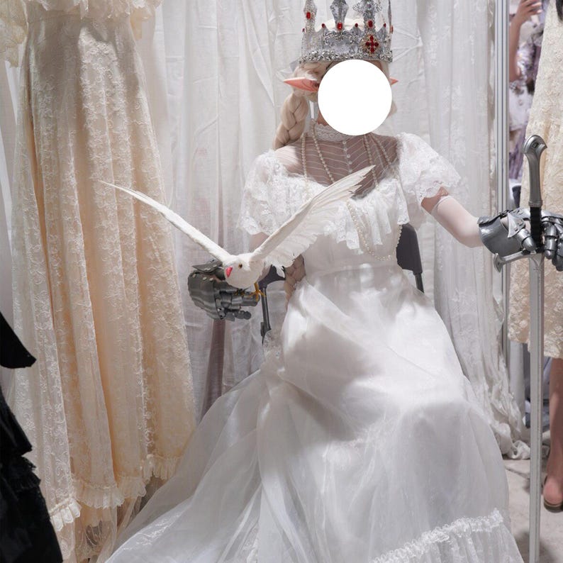 May include: A person in a white dress with a silver crown and a white dove. The dress has lace details and a layered skirt. The person is wearing a pearl necklace and has prosthetic hands. The dove is in flight.
