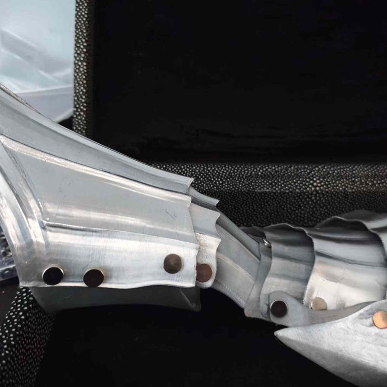May include: Close-up of articulated metal armor, likely a gauntlet or arm piece. The silver-colored metal is segmented and riveted together with small, dark rivets. The piece is set against a dark, textured background.
