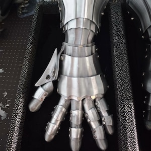 May include: A polished steel gauntlet featuring articulated metal plates covering the fingers, thumb, and wrist. The armor is constructed with multiple overlapping segments secured by small metal rivets, designed to protect the hand and forearm.