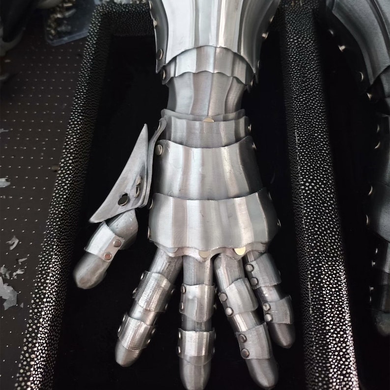 May include: A polished steel gauntlet featuring articulated metal plates covering the fingers, thumb, and wrist. The armor is constructed with multiple overlapping segments secured by small metal rivets, designed to protect the hand and forearm.