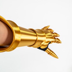 Puede incluir: Un guantelete de metal dorado que cubre el antebrazo y la mano, con segmentos de dedos articulados y puntas c&oacute;nicas. La pieza de armadura tiene un acabado liso y reflectante, con detalles de tela negra en la palma y en las articulaciones.
