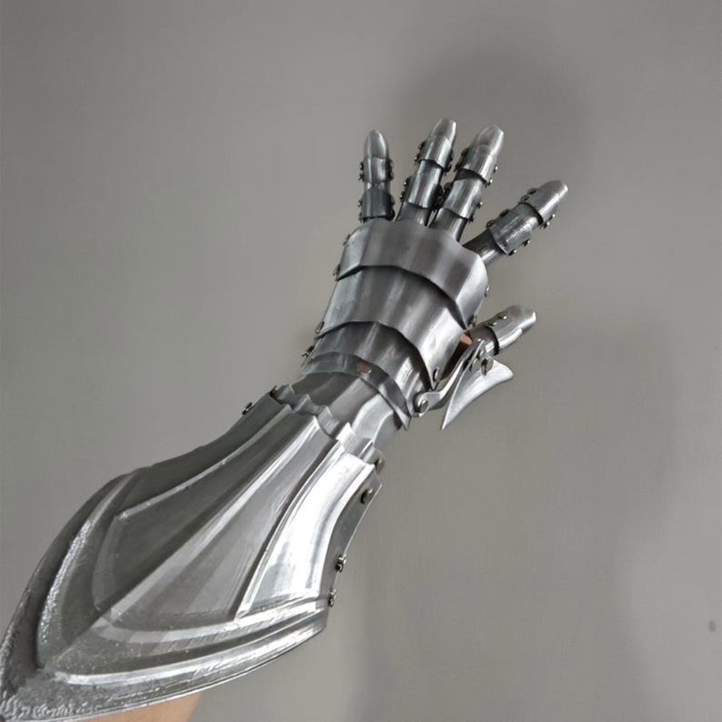 May include: A silver-colored, articulated arm and hand armor, likely for cosplay or historical reenactment. The armor is made of overlapping metal plates, with detailed finger and wrist protection. The design suggests a medieval or fantasy theme.