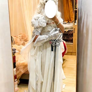 May include: A white, layered gown with a decorative bodice and flowing sleeves. The costume includes a silver gauntlet and a rapier. A white wig completes the ensemble, creating a theatrical or historical look.