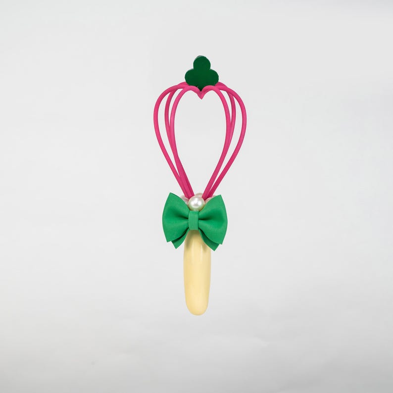 K&ouml;nnte beinhalten: Ein skurriler Zauberstab mit einem herzf&ouml;rmigen Design in Rosa, gekr&ouml;nt von einem gr&uuml;nen Kleeblatt. Eine gr&uuml;ne Schleife mit Perlenakzent sitzt &uuml;ber einem hellgelben Griff. Der Zauberstab ist vor einem wei&szlig;en Hintergrund platziert.